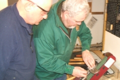 David sharpening a tool on the belt sharpener, being observed by some of the members.